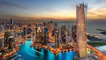 A stunning aerial view of a cityscape at dusk, showing tall, brightly lit skyscrapers surrounding a winding body of water. The sky glows with warm hues of a setting sun, and the city lights reflect off the water, creating a vibrant and dynamic scene—perfect for establishing a company in the Ras Al Khaimah Free Trade Zone.